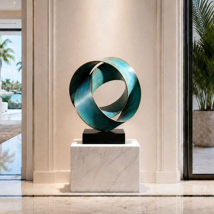 Circular bronze ribbon sculpture with blue-green patina displayed on a white stone pedestal in a bright interior