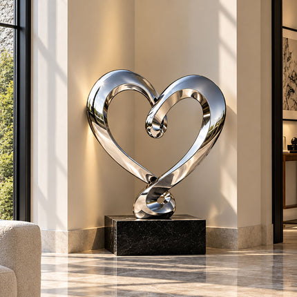 Stainless steel heart sculpture on a low black base in a landscaped courtyard beside a modern building.