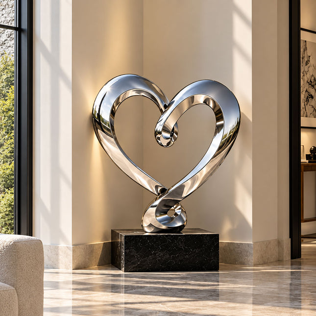 Stainless steel heart sculpture on a low black base in a landscaped courtyard beside a modern building.