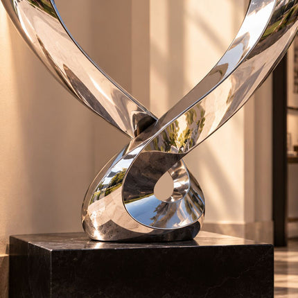 Close-up of the crossed metal bands and lower loop of a stainless steel heart sculpture above a dark base.