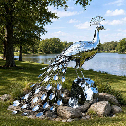 Mirror polished stainless steel peacock sculpture with a long trailing tail displayed on a grassy lakeside landscape
